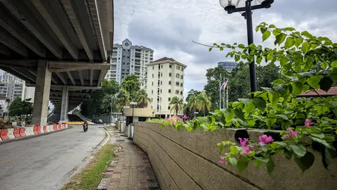 Wickham Manor: A street-level view from under a highway overpass, showing a road with a motorcyclist, a sidewalk, and a beige apartment building in the background.