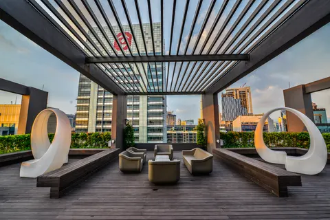 The Manhattan: A furnished outdoor lounge area on a rooftop deck, featuring wooden flooring, seating, and modern white sculptures under a pergola.