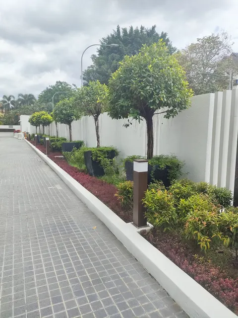 Residensi R8, Ampang Hilir: A paved walkway lined with a manicaped garden bed featuring small trees, shrubs, and modern bollard lights, running alongside a white boundary wall.