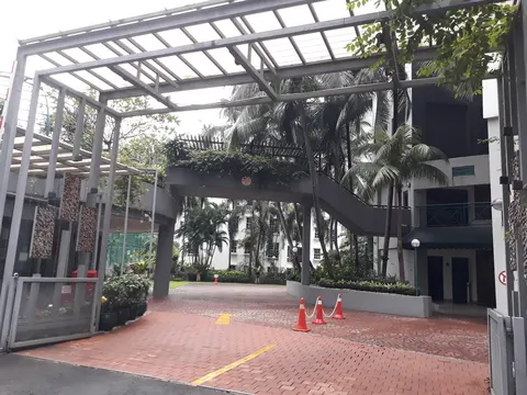 D'Mayang: An outdoor view of a development entrance featuring a steel pergola, paved driveway with red cones, landscaped garden, and a building with a dark facade.