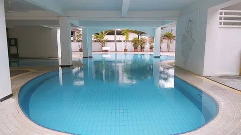 The Pines: An indoor swimming pool with blue tiles and water, surrounded by white pillars and a tiled deck, with potted plants and an outdoor area visible through an opening.