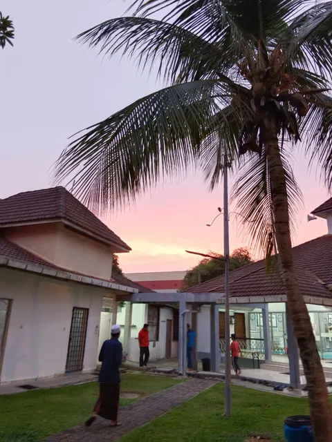 Gugusan Dedap: Residents walk through the landscaped grounds of a residential complex with shingled roof buildings at dusk, with a palm tree in the foreground.