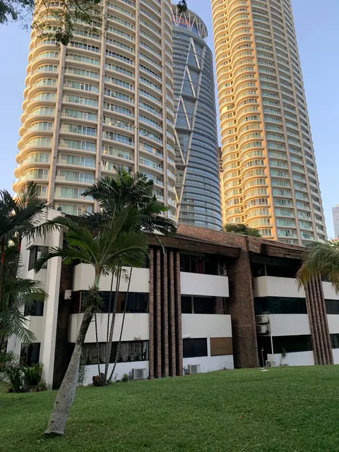 Desa Kudalari: A view of the exterior of a residential development, featuring tall towers with curved balconades and a low-rise building with brick and white facade elements in the foreground, set against a lawn with palm trees.