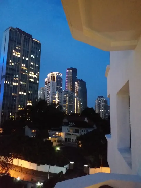 Sri Kia Peng: A nighttime view from a balcony looking out over trees and houses towards a city skyline with illuminated high-rise buildings.