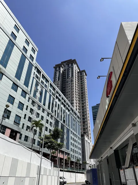 Menara Avenue: A low-angle street view showing the facade of a commercial building on the left and the entrance canopy of a shopping mall on the right, with a high-rise residential tower visible in the background against a clear blue sky.
