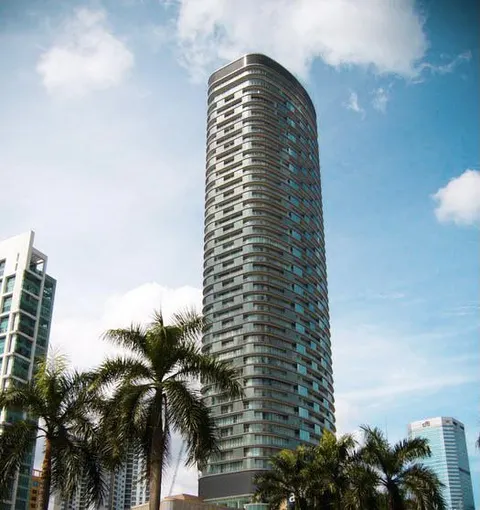 K Residence: A finished, cylindrical high-rise tower with a dark glass facade stands against a blue sky, framed by palm trees and adjacent buildings.