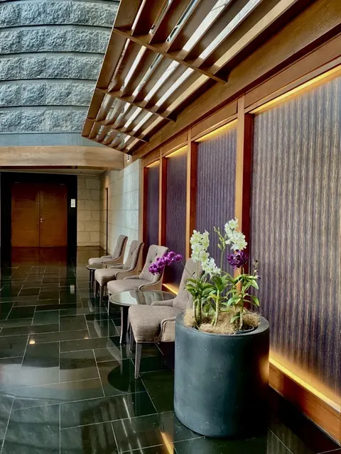 The Binjai On The Park: A sophisticated hotel or corporate lobby lounge featuring dark polished floors, a row of armchairs, and a large floral arrangement of white and purple orchids, set beneath a wooden slatted ceiling.