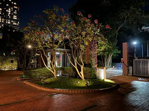 Dedaun Condominium: A night view of a landscaped entrance area for a residence, featuring a flowering tree, a security booth, and a gate, with a high-rise building in the background.