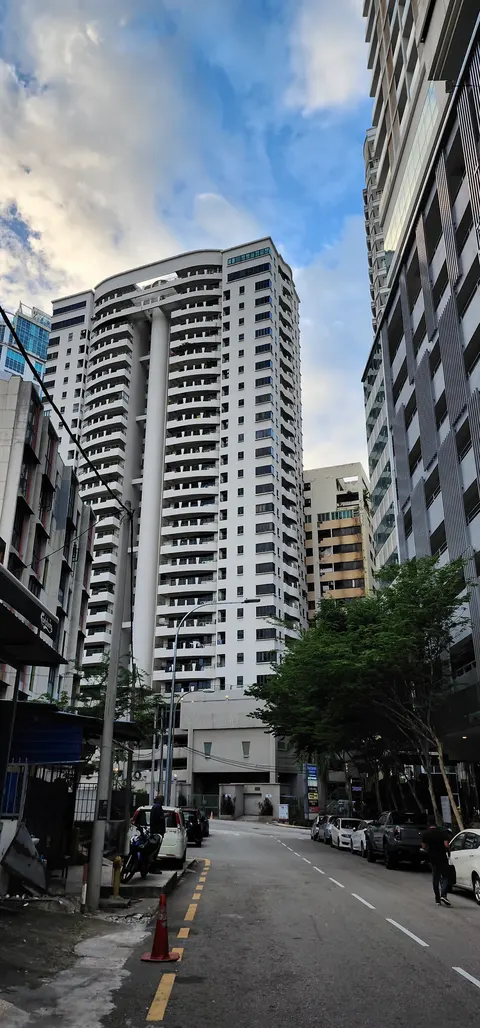 Seri Bukit Ceylon: A tall white residential building with vertical pillars and many windows is viewed from a street, surrounded by other urban buildings and parked cars.