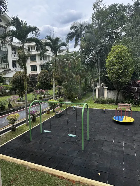Regal Villa: A children's playground area with a swing set, a spinner, and a bench, paved with black rubber safety tiles and surrounded by a landscaped garden with palm trees.