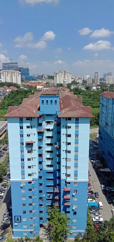 Pangsapuri Sri Rakyat: A high-angle exterior shot of a tall, blue residential building with a reddish-brown tiled roof and external staircases, set against a backdrop of other apartment complexes and a blue sky.