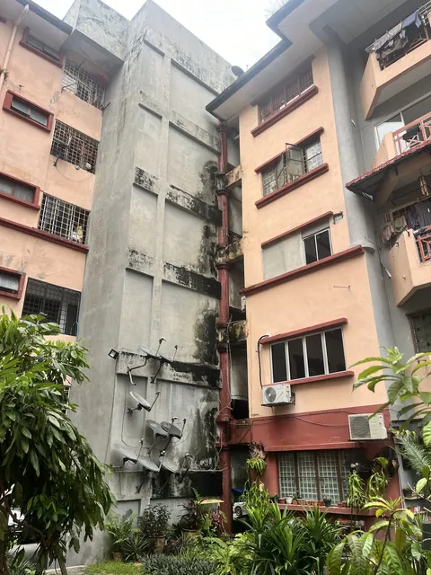 Pangsapuri Kayangan: The weathered concrete facade and pink exterior wall of a housing block, featuring multiple satellite dishes, air conditioning units, and ground-level tropical foliage.