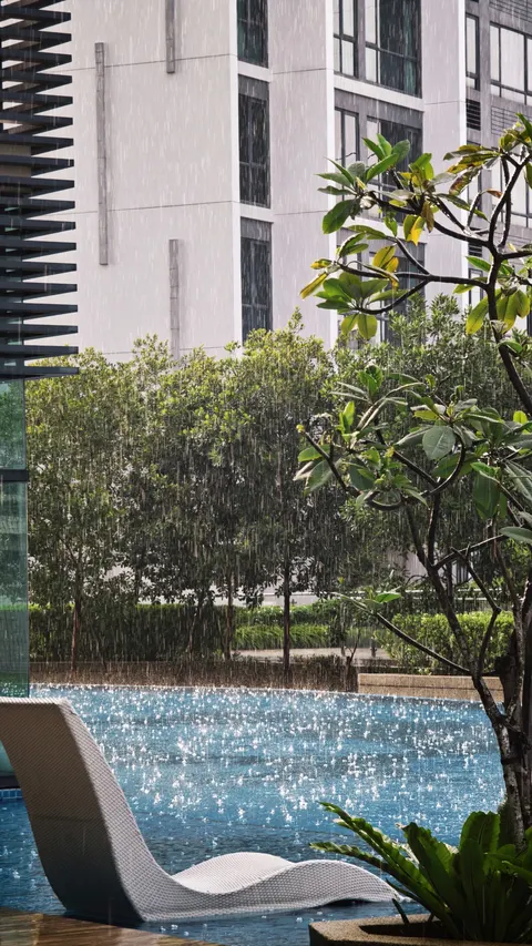 Greenfield Residence: A view of a swimming pool with a lounge chair during a rainstorm, with green trees and a residential building in the background.