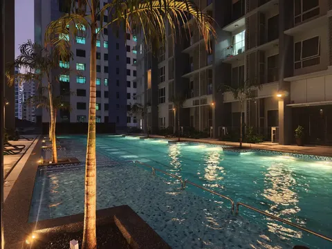 M-Suites @ Embassy Row: An outdoor swimming pool and leisure area at night, featuring palm trees, lounge chairs, and apartment towers in the background.