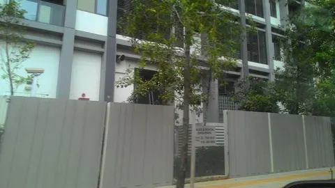 Katana II: The exterior of a residential development, viewed from across the street, showing its facade, entrance gate, and surrounding trees.