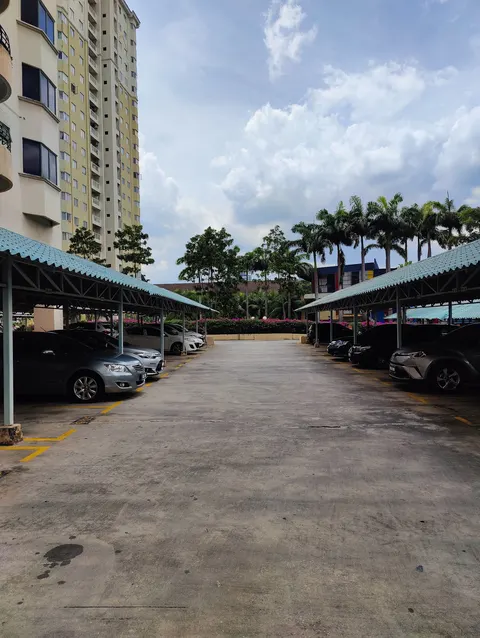 Duta Ria: An outdoor car park with covered parking bays, parked cars, and a high-rise residential building in the background.