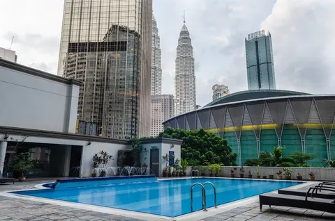 Menara Pinang: A rooftop swimming pool with sun loungers, viewed against a backdrop of the Petronas Towers and a large arena-like building.