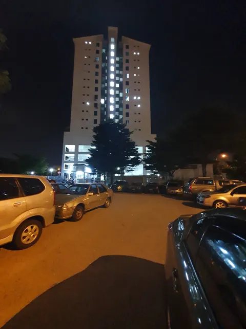 Kasturi Idaman: A tall residential building at night, illuminated from within and below, with a parking lot full of cars in the foreground.