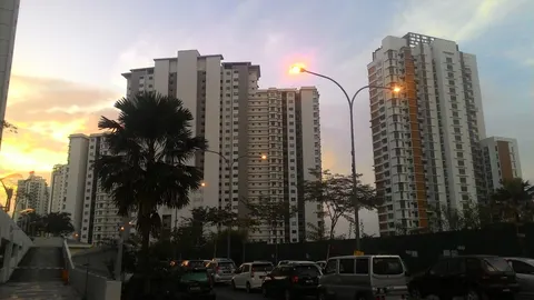 Residensi Laguna: Several high-rise residential buildings with a palm tree and cars in the foreground at sunset.