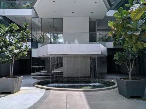 Cascades Residency: A courtyard with a circular water feature and a waterfall flowing from a concrete balcony, flanked by potted fiddle-leaf fig trees.