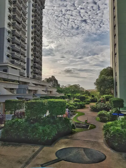 Vista Komanwel B: A view of a landscaped pathway and manicured gardens within a residential complex, flanked by high-rise apartment buildings.