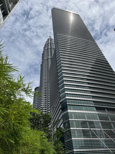One KL: A low-angle view looking up at the facade of a modern glass skyscraper, with the Petronas Twin Towers visible in the background under a cloudy sky.