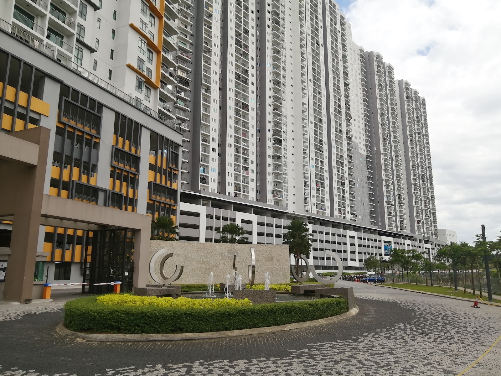 Parkhill Residence Bukit Jalil: A view of the exterior entrance and high-rise residential towers of a development, featuring a paved driveway, a landscaped planter with a fountain, and a parking structure in the background.