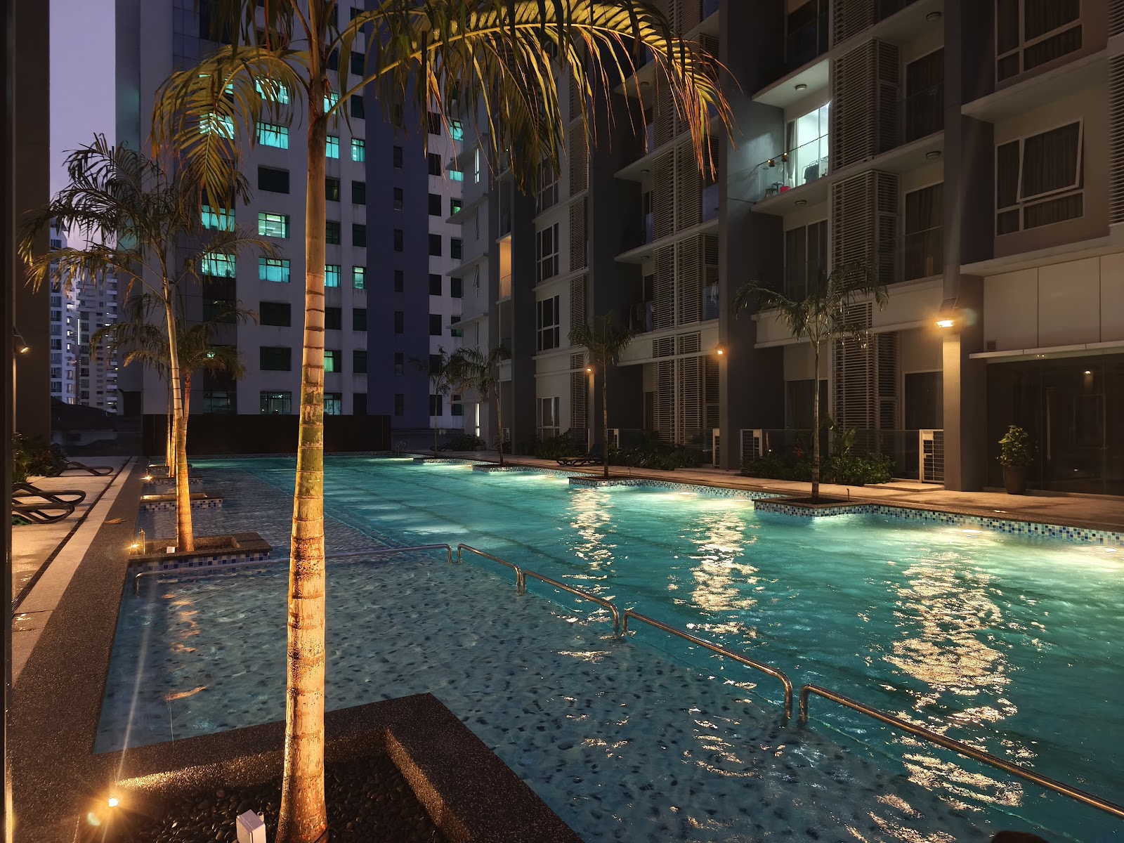 M-Suites @ Embassy Row: An outdoor swimming pool and leisure area at night, featuring palm trees, lounge chairs, and apartment towers in the background.