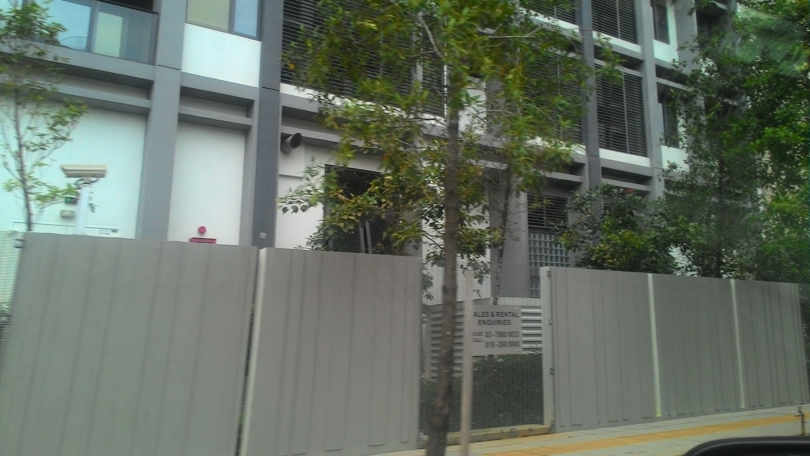 Katana II: The exterior of a residential development, viewed from across the street, showing its facade, entrance gate, and surrounding trees.