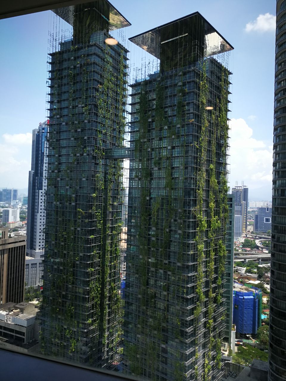Le Nouvel KLCC: A high-angle view from a window looking out onto the green-covered towers of Maxis @ The Vertical in Kuala Lumpur.
