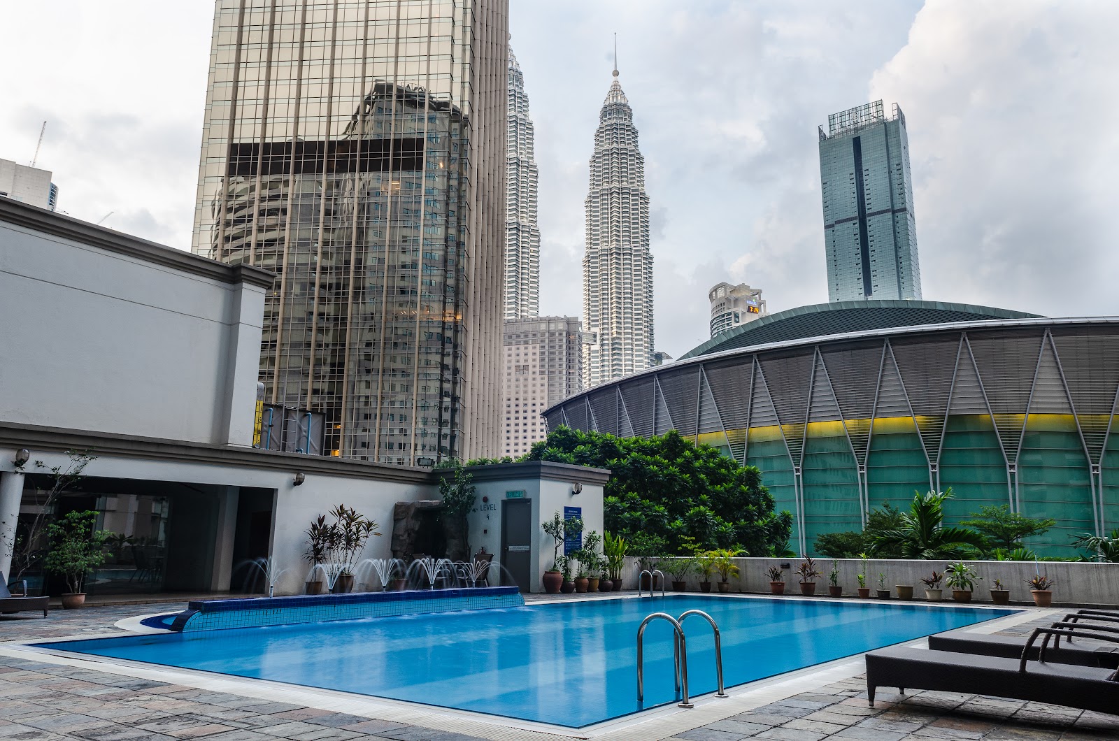 Menara Pinang: A rooftop swimming pool with sun loungers, viewed against a backdrop of the Petronas Towers and a large arena-like building.