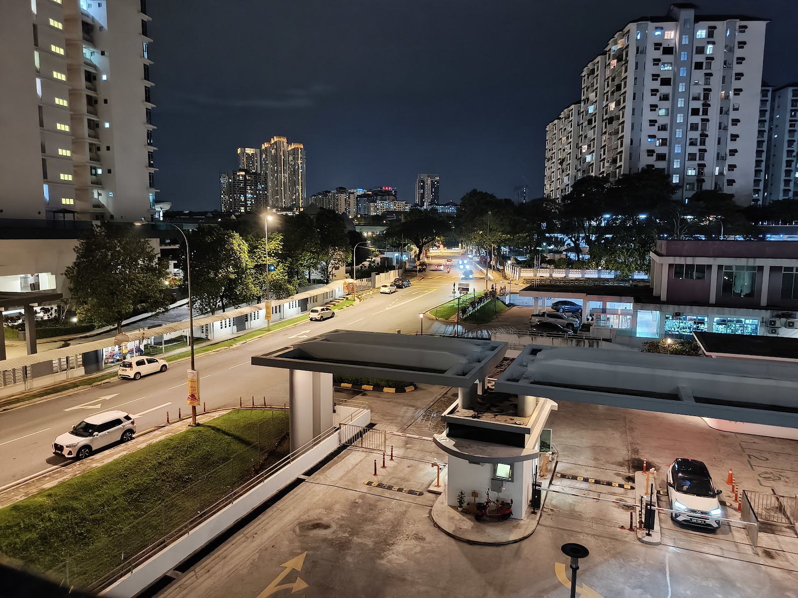 Kiara Residence 1: A night view from an elevated position overlooking a car park entrance and a main road in a residential area with tall apartment buildings in the background.