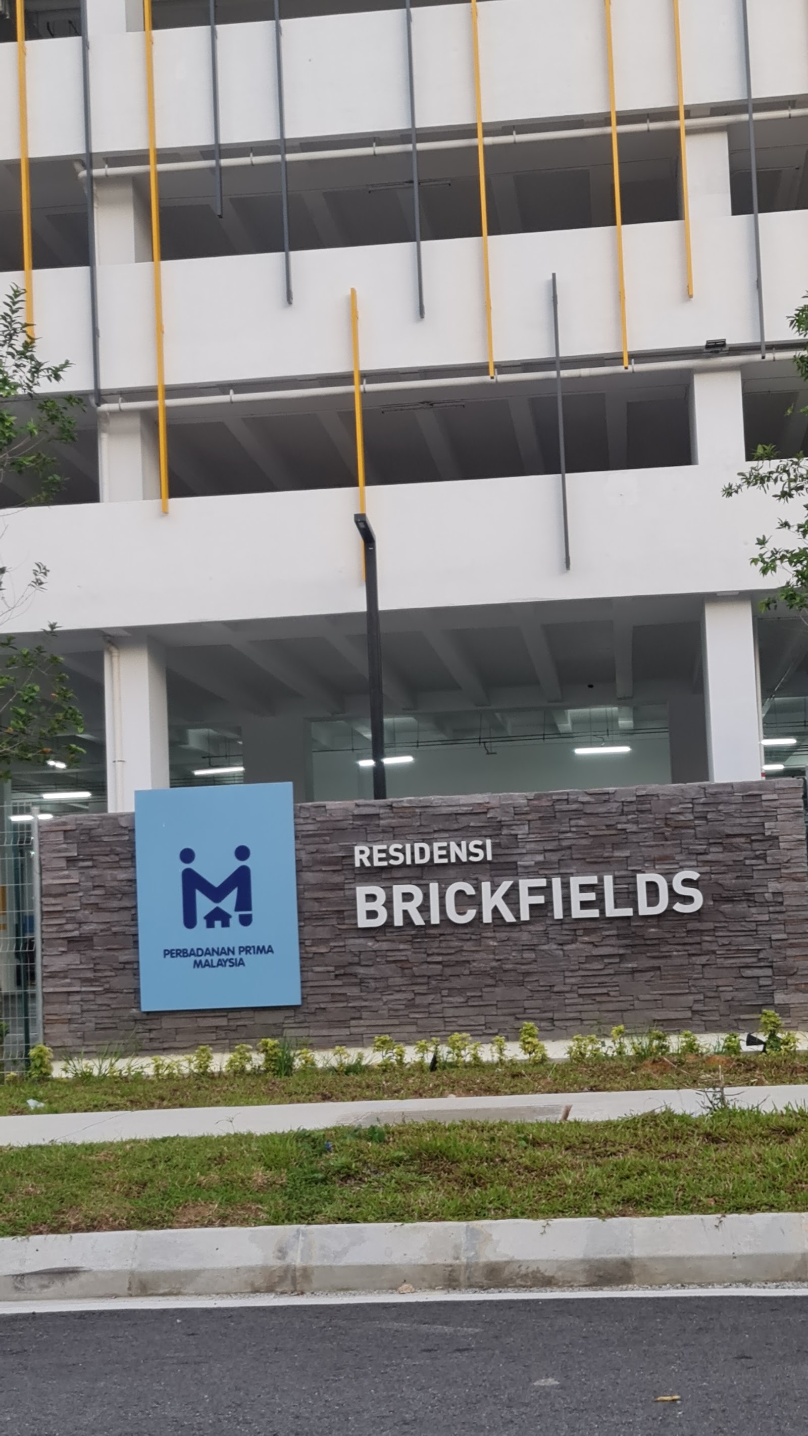 RESIDENSI BRICKFIELDS: A view of the entrance signage for Residensi Brickfields featuring a blue corporate sign and white letters on a stone wall, set against a multi-story parking structure.
