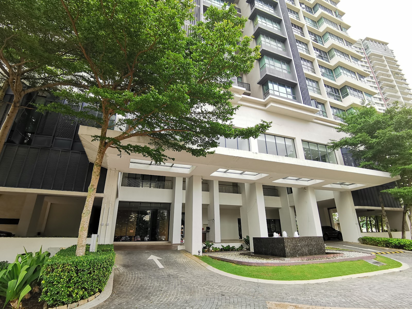 KM1: Entrance canopy of a residential building with paved driveway, landscaping, and high-rise tower facade in the background.