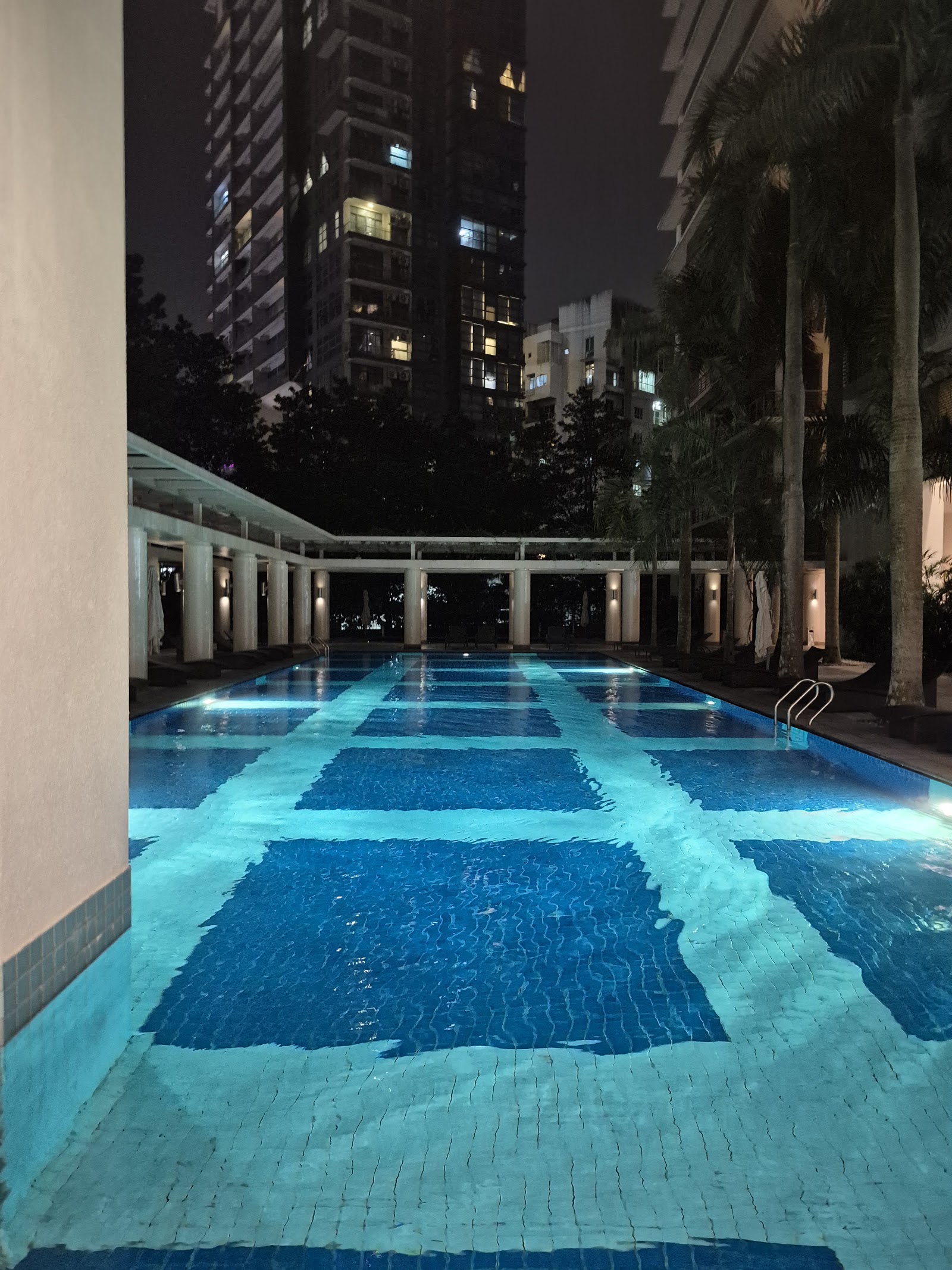 Hampshire Park: An illuminated outdoor swimming pool at night, surrounded by a colonnade and palm trees, with a high-rise building in the background.