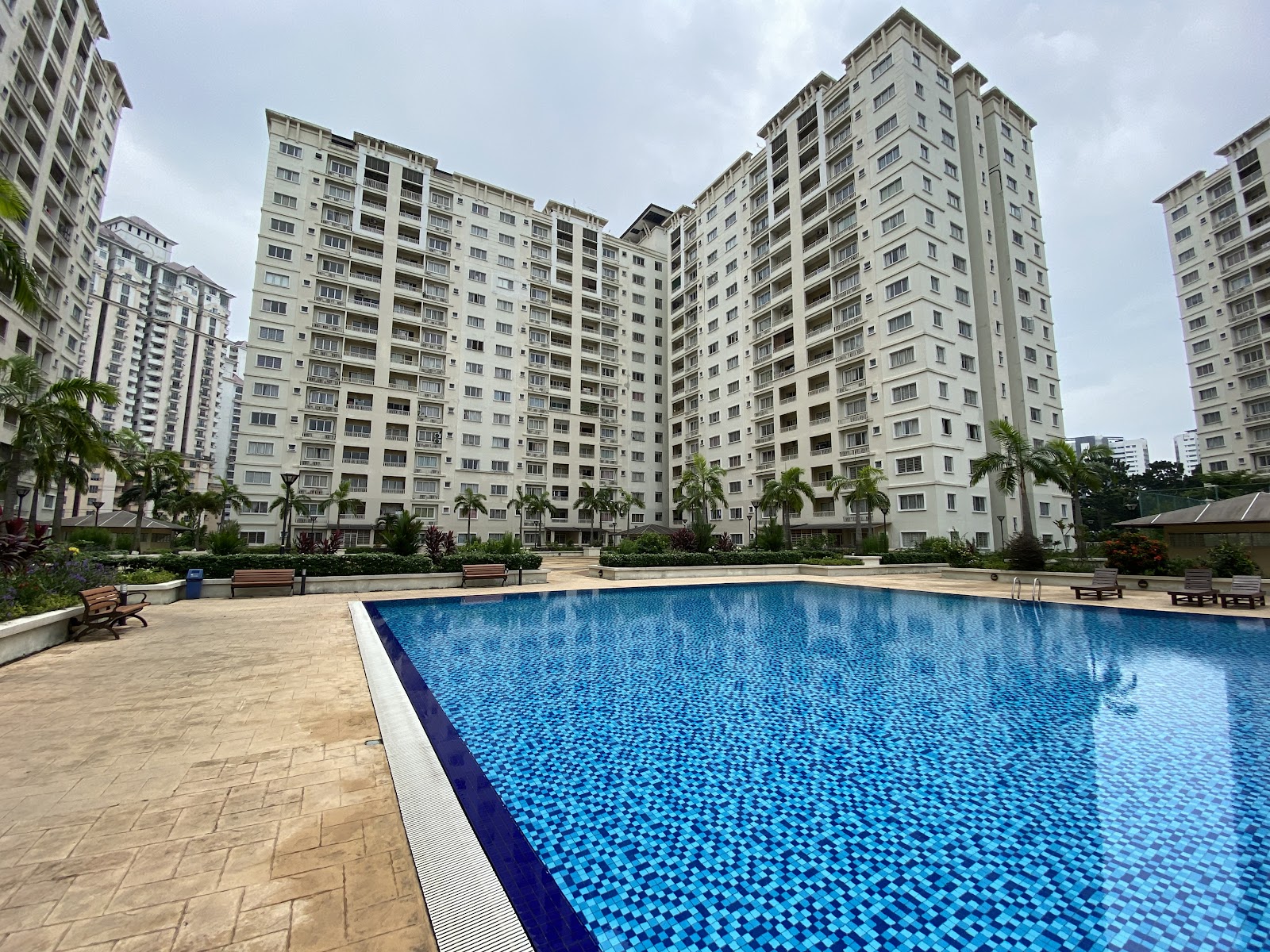 Suriamas: A large residential swimming pool with blue mosaic tiles and surrounding paved deck with benches, set in a garden landscape with tall apartment towers in the background.