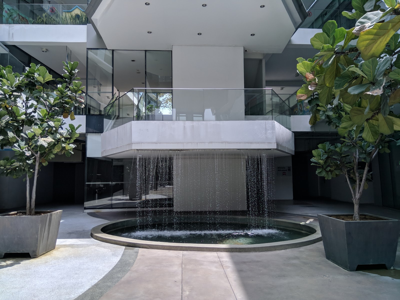 Cascades Residency: A courtyard with a circular water feature and a waterfall flowing from a concrete balcony, flanked by potted fiddle-leaf fig trees.