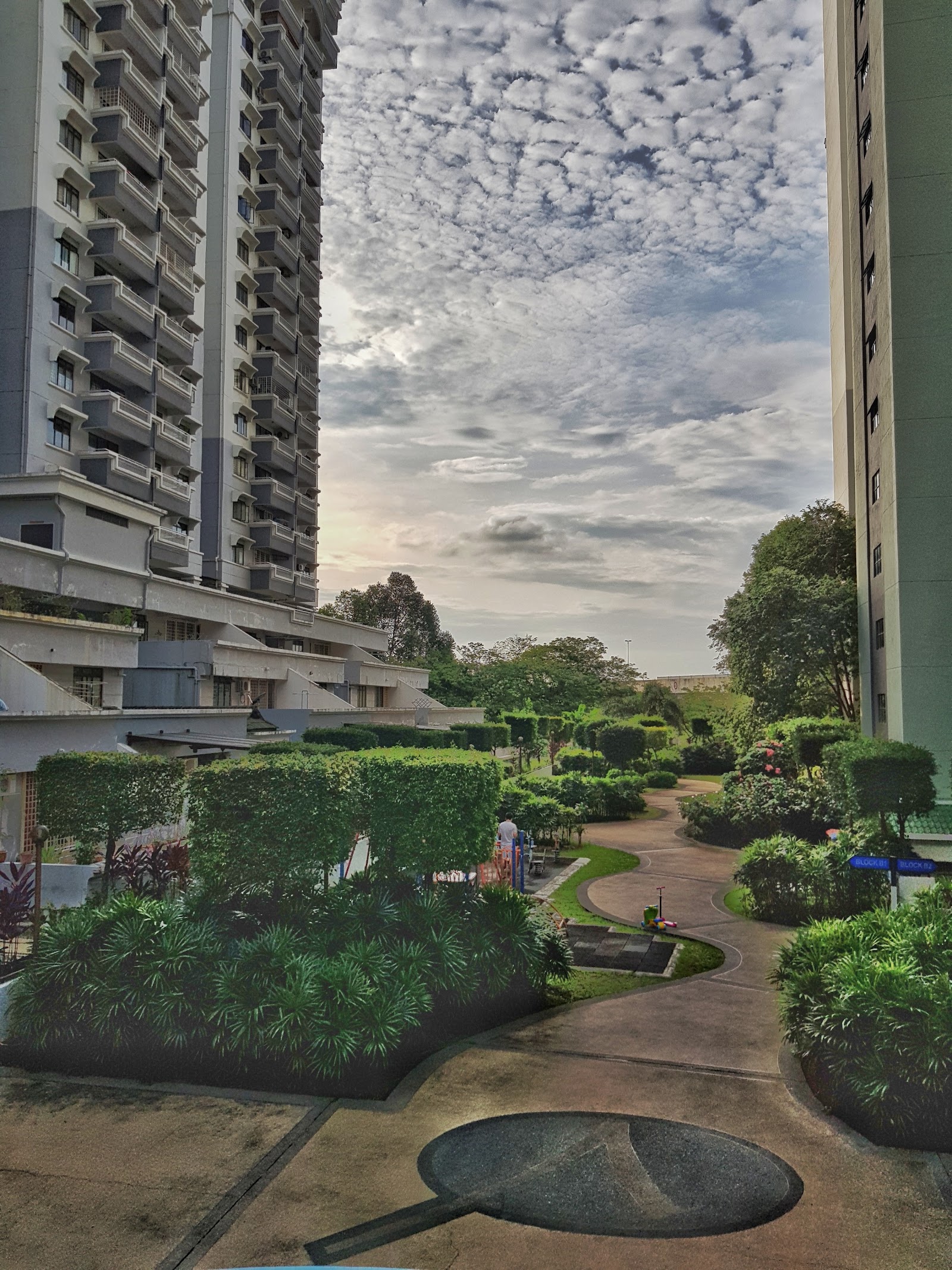 Vista Komanwel B: A view of a landscaped pathway and manicured gardens within a residential complex, flanked by high-rise apartment buildings.