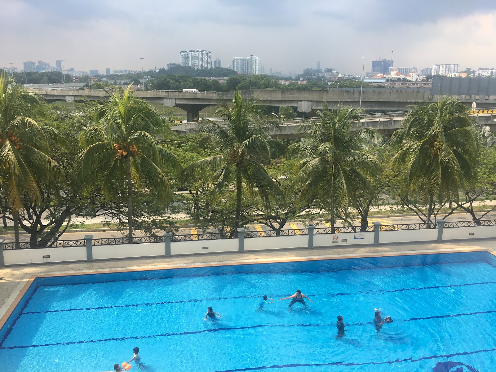 Vista Komanwel: A swimming pool with several people swimming, located on a high floor, with a view of palm trees and a highway in the background.