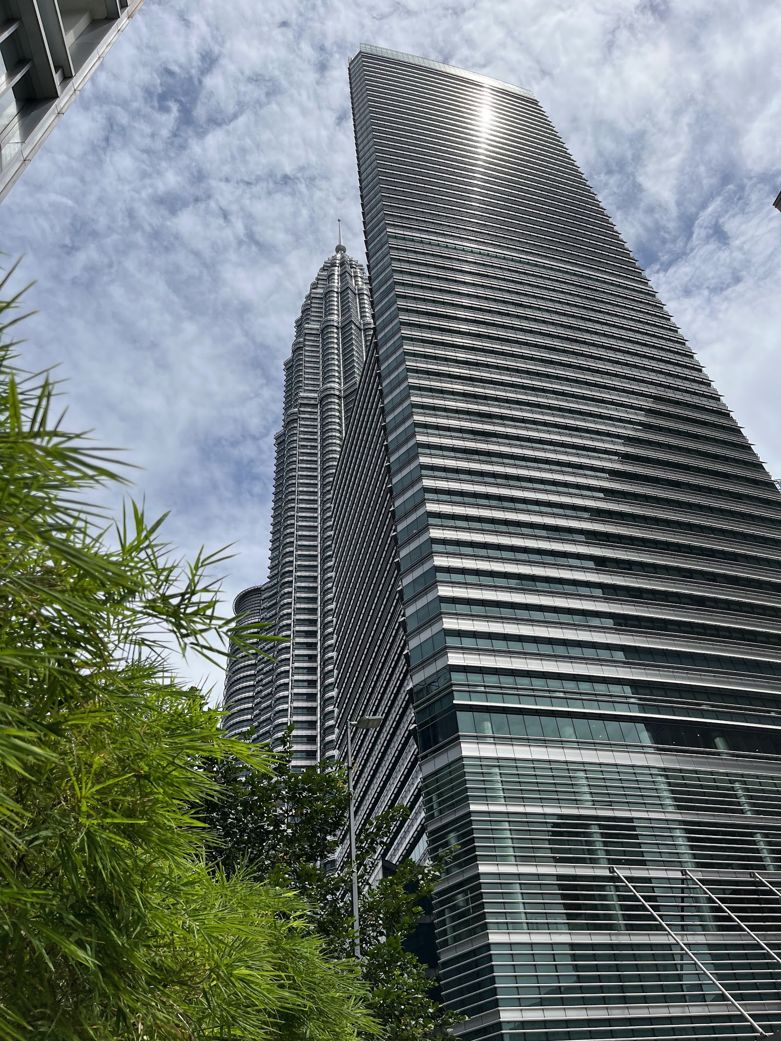 One KL: A low-angle view looking up at the facade of a modern glass skyscraper, with the Petronas Twin Towers visible in the background under a cloudy sky.